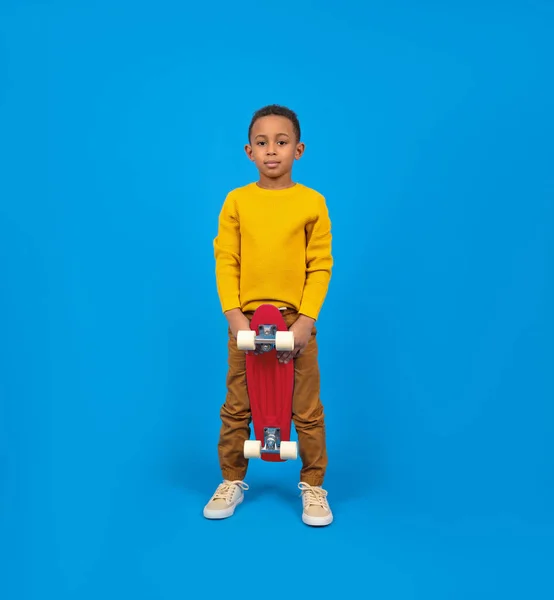 Cute African-American teenager in casual clothes, yellow sweater and brown pants, standing and holding red skateboard and smiling against blue background. Concept of activity and happy childhood. — Stock Photo, Image