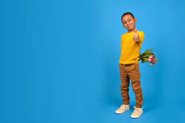 African-American boy with bouquet of tulips hides behind his back, finger-up gesture of approval or like on blue background. concept mothers Day, fathers day, Valentines day gift for woman — Stock Photo, Image