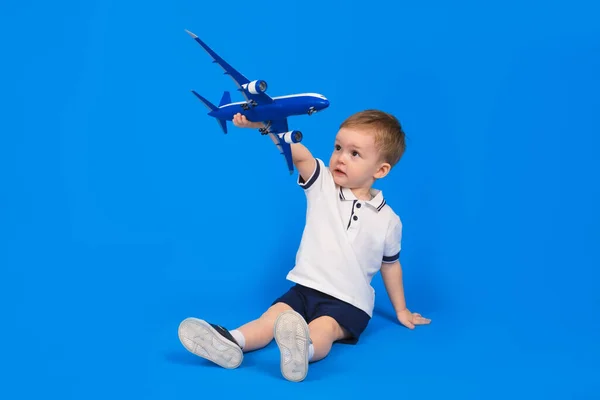 Happy child sits holding plane in his hand and imagining how he is flying on plane on blue background. Imagination, creativity and concept ideas. — Stock Photo, Image