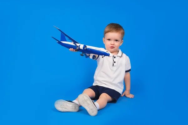 Criança feliz senta-se segurando avião em sua mão e imaginando como ele está voando no avião em fundo azul. Imaginação, criatividade e ideias conceituais. — Fotografia de Stock