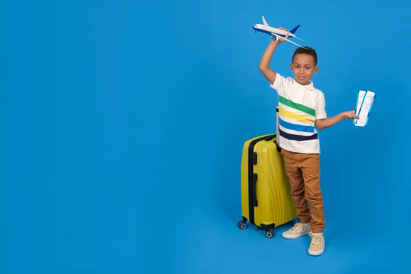 Chico viajero afroamericano está listo para el viaje, mostrando pasaporte y billetes de avión es feliz, bolsa de viaje amarilla, tiene una oferta especial de una empresa de viajes, sobre un fondo azul. — Foto de Stock