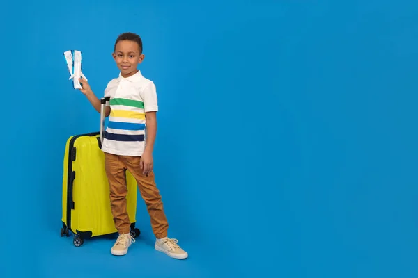Chico viajero afroamericano está listo para el viaje, mostrando pasaporte y billetes de avión es feliz, bolsa de viaje amarilla, tiene una oferta especial de una empresa de viajes, sobre un fondo azul. — Foto de Stock