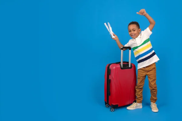 Viajero turista chico afroamericano sobre fondo azul. Pasajero viajando al extranjero durante el fin de semana. Vuelos al viaje a pie fuera de la maleta para guardar pasaporte, tarjeta de embarque, billetes de avión — Foto de Stock