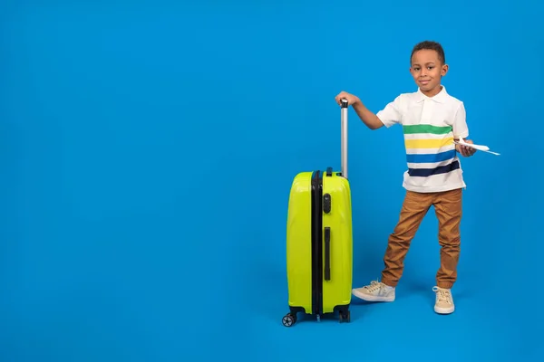 Chico viajero afroamericano está listo para el viaje, mostrando pasaporte y billetes de avión es feliz, bolsa de viaje amarilla, tiene una oferta especial de una empresa de viajes, sobre un fondo azul. — Foto de Stock