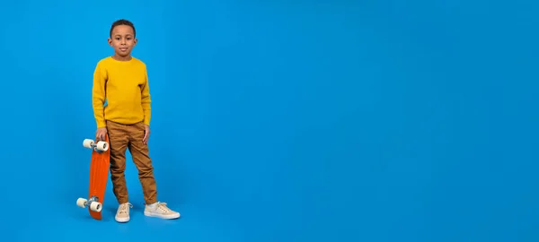 カジュアルな服、黄色のセーターと茶色のパンツでかわいいアフリカ系アメリカ人のティーンエイジャーは、立って、赤いスケートボードを保持し、青い背景に笑顔。活動と幸せな子供時代の概念. — ストック写真