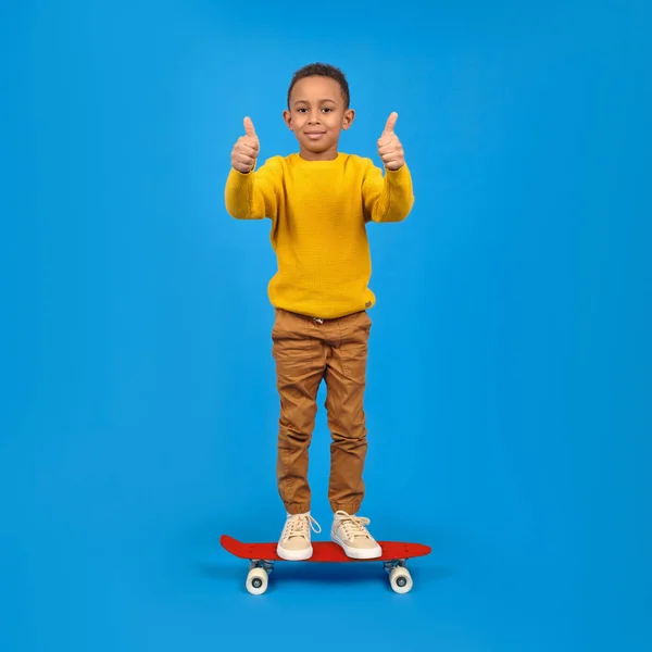 Happy African American boy in casual clothes posing with a skateboard and giving a thumbs up to the camera on a blue background. Concept of activity and happy childhood. — Stock Photo, Image