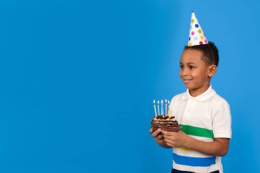 Kafasında küçük bir parti konisi olan mutlu Afrikalı Amerikalı çocuk ellerinde mumlarla çikolatalı pasta tutarak ve mavi arka planda bayramda sevinerek doğum gününü kutluyor. Doğum günü kavramı