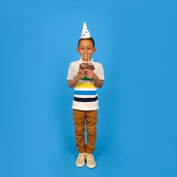 Feliz niño afroamericano celebra su cumpleaños sosteniendo pastel de chocolate en las manos con velas encendidas y regocijándose en las vacaciones sobre fondo azul. Concepto de fiesta de cumpleaños, espacio para copiar — Foto de Stock