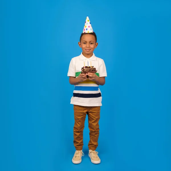 Happy African American boy with funny little party cone on head celebrates birthday holding chocolate cake in hands with burning candles and rejoicing in holiday on blue background. Birthday concept — Stock Photo, Image