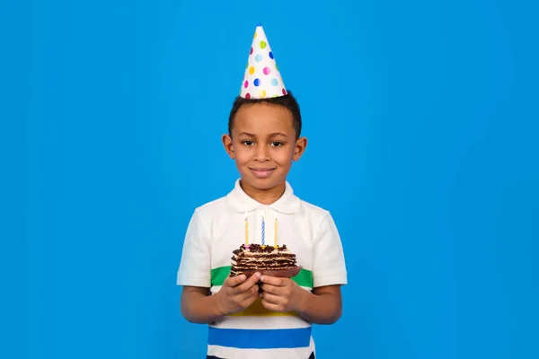 Feliz niño afroamericano celebra su cumpleaños sosteniendo pastel de chocolate en las manos con velas encendidas y regocijándose en las vacaciones sobre fondo azul. Concepto de fiesta de cumpleaños, espacio para copiar — Foto de Stock