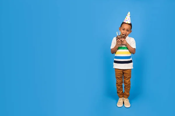 Boldog afro-amerikai fiú egy vicces kis party kúp a fején ünnepli születésnapját kezében csokoládé torta és harapás torta örül a kék háttérben. Születésnapi party koncepció — Stock Fotó