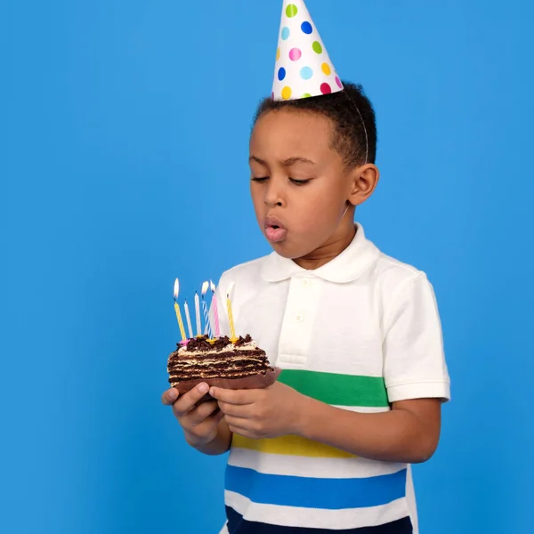 African-American boy blows out emotionally candles on birthday cake, wearing white polo and festive hat on his head standing on blue studio background. holiday and birthday party with place for text — Stock Photo, Image