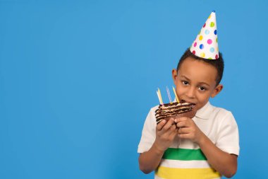 Kafasında küçük bir parti konisi olan mutlu Afro-Amerikalı çocuk ellerinde çikolatalı pasta tutarak ve mavi arka planda pasta yiyerek doğum gününü kutluyor. Doğum günü partisi konsepti