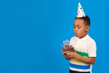 Afro-Amerikalı çocuk duygusal olarak pastaya mum üflüyor, beyaz polo giyiyor ve kafasında şenlik şapkası var mavi stüdyo arka planında duruyor. tatil ve doğum günü partisi için metin yeri 