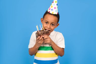 Kafasında küçük bir parti konisi olan mutlu Afro-Amerikalı çocuk ellerinde çikolatalı pasta tutarak ve mavi arka planda pasta yiyerek doğum gününü kutluyor. Doğum günü partisi konsepti