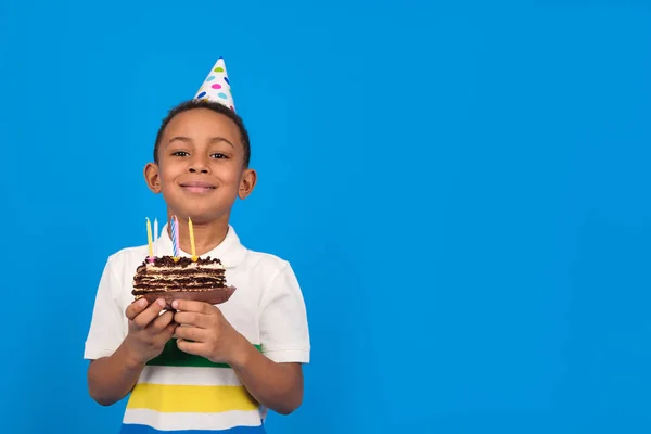 Happy funny cute African American boy with funny little party cone on his head holding cake and smiling blue background, his look expresses pure joy and happiness. Birthday party concept, copy space — Stock Photo, Image
