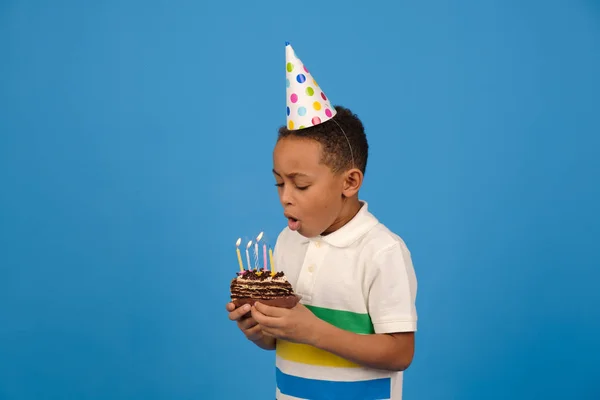 African-American boy blows out emotionally candles on birthday cake, wearing white polo and festive hat on his head standing on blue studio background. holiday and birthday party with place for text — Stock Photo, Image