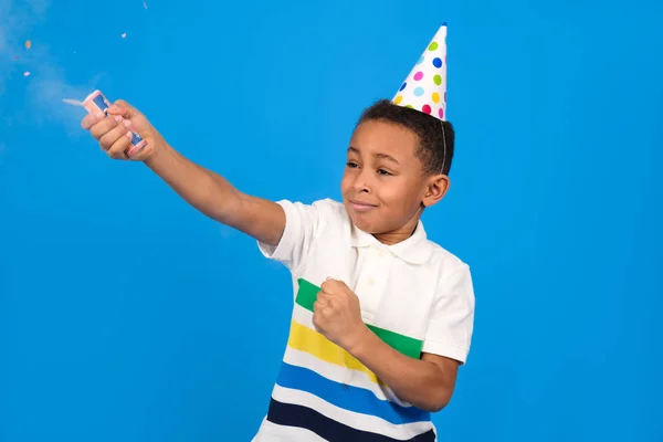 Rapaz afro-americano explode flapper segurando-a em sua mão vestida com pólo branco e boné de festa fica em fundo estúdio azul. Conceito de aniversário e feriados festa conceito com lugar para texto . — Fotografia de Stock