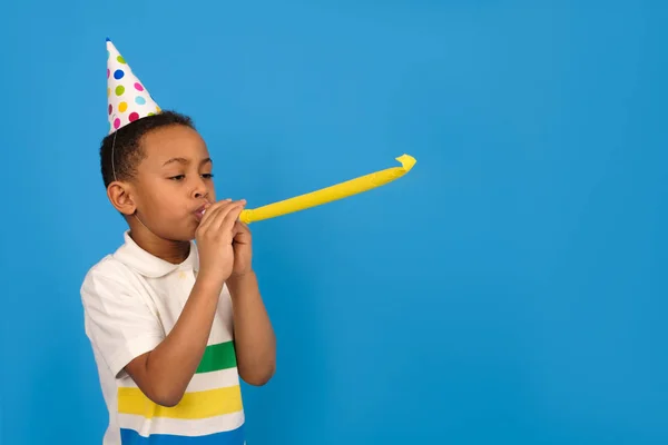 Feliz niño afroamericano celebra su cumpleaños haciendo sonar un silbato sobre un fondo azul. Concepto de vacaciones y fiesta con espacio para el texto . — Foto de Stock