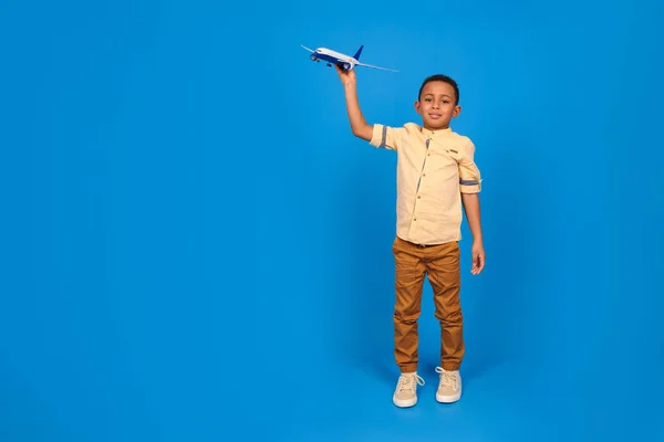 茶色のパンツを着たアフリカ系アメリカ人の幸せな少年と、おもちゃの飛行機で遊ぶライトシャツは、青い背景を背景に飛行機で飛ぶことを想像しています。家族旅行や観光、夏休みの結果. — ストック写真