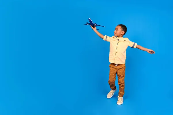 幸せなアフリカ系アメリカ人の少年はおもちゃの飛行機で遊び、手を脇に置き、青い背景を背景に飛行機に乗っている自分を想像します。家族旅行や観光、夏休みのコンセプト. — ストック写真