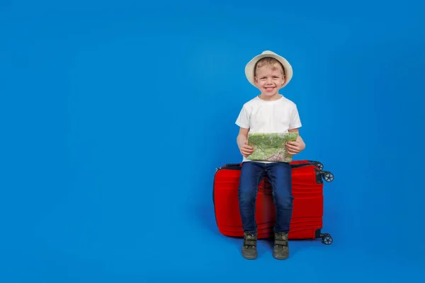 Niño turista con una tarjeta de viaje se sienta en una maleta roja para viajar y se mira en un mapa, fondo azul. Un concepto para el turismo con lugar para el texto . — Foto de Stock