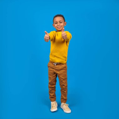 Mutlu Afrikalı genç Afrikalı Amerikalı jest gibi başparmak kaldırıyor, kameraya bakın, gülümseyen çocuk, memnun müşteri iyi hizmet öneriyor, mavi stüdyo arka planında izole edilmiş.