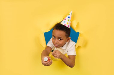Beyaz polo tişörtlü ve tatil şapkalı mutlu Afro-Amerikan çocuk sarı arka planda yırtık kağıt kenarlı bir alçı tahtasını havaya uçuruyor. Doğum günü ya da Yeni Yıl Arifesi, kutlama konsepti.