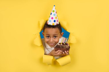 Mutlu Afro-Amerikalı çocuk doğum gününü elinde mumlarla çikolatalı pasta tutarak ve sarı yırtık kağıtta tatilin tadını çıkararak kutluyor. Konsept parti doğum günü, kopya için boşluk