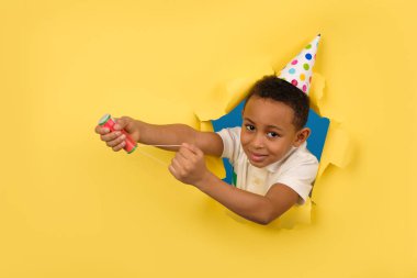 Beyaz polo tişörtlü ve tatil şapkalı mutlu Afro-Amerikan çocuk sarı arka planda yırtık kağıt kenarlı bir alçı tahtasını havaya uçuruyor. Doğum günü ya da Yeni Yıl Arifesi, kutlama konsepti.