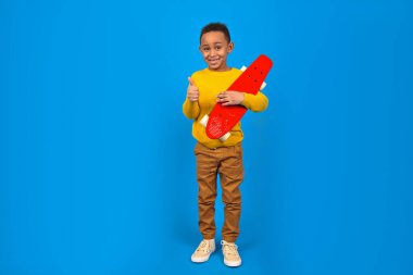 Sarı kazaklı, kahverengi pantolonlu, kırmızı kaykaylı ve mavi arka planda gülümseyen mutlu Afro-Amerikalı ergen. Aktivite ve mutlu çocukluk kavramı.