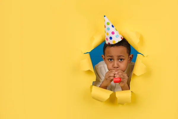 Boldog afro-amerikai fiú fújja ünnepi pipa születésnapi party egy kúp sapka a fején egy sárga szakadt háttér szakadt élek papír. A gyerekek szórakoztatóak. A kölyök szülinapi bulit ünnepel.. — Stock Fotó