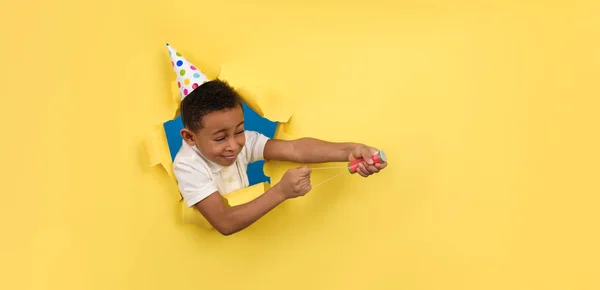 Boldog afro-amerikai fiú fehér pólóban és ünnepi sapkában felrobbant egy clapperboard a kezében egy sárga stúdió háttérben szakadt papírszélekkel. Születésnap vagy Szilveszter, ünnepi koncepció. — Stock Fotó