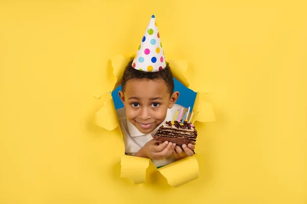 Menino afro-americano feliz comemora o aniversário segurando um bolo de chocolate em suas mãos com velas e desfrutando de férias em fundo de papel rasgado amarelo. Conceito festa aniversário, espaço para cópia — Fotografia de Stock