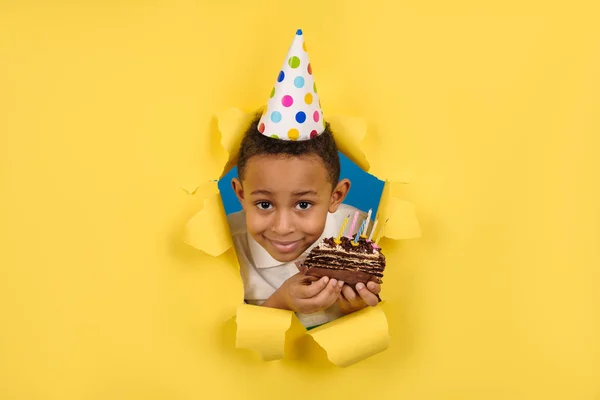 Feliz niño afroamericano celebra su cumpleaños sosteniendo un pastel de chocolate en sus manos con velas y disfrutando de las vacaciones en un fondo de papel amarillo roto. Concepto fiesta cumpleaños, espacio para la copia — Foto de Stock