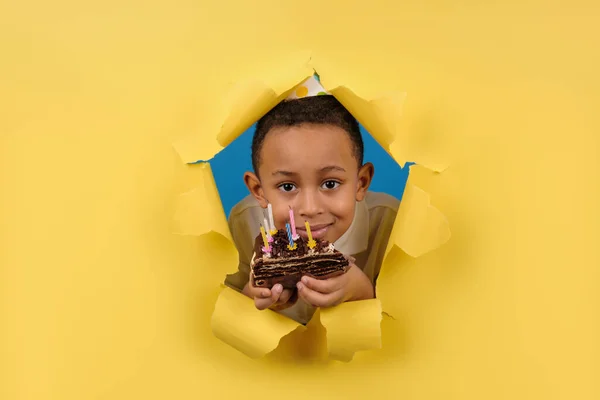 Feliz niño afroamericano celebra su cumpleaños sosteniendo un pastel de chocolate en sus manos con velas y disfrutando de las vacaciones en un fondo de papel amarillo roto. Concepto fiesta cumpleaños, espacio para la copia — Foto de Stock