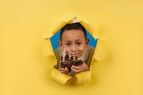 Menino afro-americano feliz comemora o aniversário segurando um bolo de chocolate em suas mãos com velas e desfrutando de férias em fundo de papel rasgado amarelo. Conceito festa aniversário, espaço para cópia — Fotografia de Stock