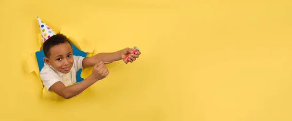 Menino americano africano feliz em camisa de pólo branco e chapéu de férias explode uma tábua de palmas em suas mãos em um fundo de estúdio amarelo com bordas rasgadas de papel. Aniversário ou Ano Novo, conceito de celebração. — Fotografia de Stock