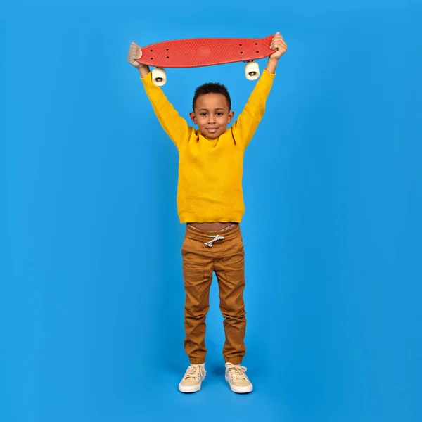 Portrait African-American teenager in casual clothes, yellow sweater and brown pants, standing and holding red skateboard and smiling against blue background. Concept of activity and happy childhood — Stock Photo, Image