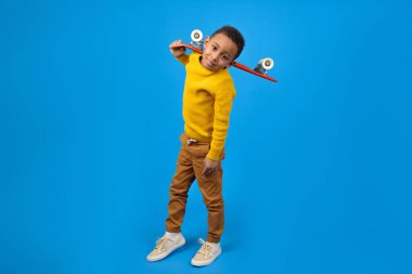 Sarı süveter ve kahverengi pantolon giyen, kırmızı kaykay tutan ve mavi stüdyo arka planında gülümseyen sevimli Afro-Amerikan ergen. Aktivite ve mutlu çocukluk kavramı