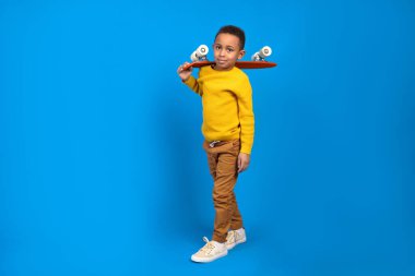 Sarı süveter ve kahverengi pantolon giyen, kırmızı kaykay tutan ve mavi stüdyo arka planında gülümseyen sevimli Afro-Amerikan ergen. Aktivite ve mutlu çocukluk kavramı