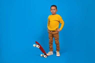 Sarı kazaklı, kahverengi pantolonlu, kırmızı kaykaylı ve mavi arka planda gülümseyen mutlu Afro-Amerikalı ergen. Aktivite ve mutlu çocukluk kavramı.