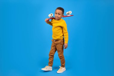 Sarı süveter ve kahverengi pantolon giyen, kırmızı kaykay tutan ve mavi stüdyo arka planında gülümseyen sevimli Afro-Amerikan ergen. Aktivite ve mutlu çocukluk kavramı