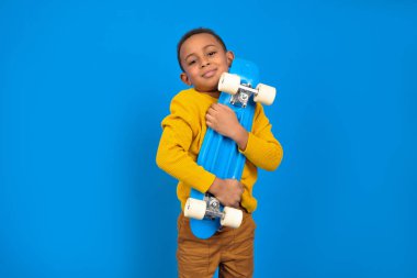 Sıradan kıyafetler içinde, sarı kazak ve kahverengi pantolon giymiş mavi kaykay tutan ve mavi arka planda gülümseyen Afro-Amerikalı bir genç portresi. Aktivite ve mutlu çocukluk kavramı.