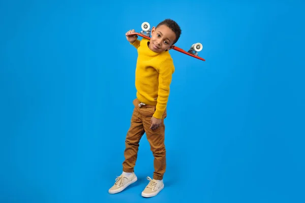 Cute African-American teenager in casual clothes yellow sweater and brown pants, walking and holding red skateboard and smiling against blue studio background. Concept of activity and happy childhood — Stock Photo, Image