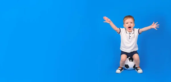 Boy sits on soccer ball and rejoices in victory, raising his arms to top against blue background. Emotions of winner and champion, place for text. Concept of winning and achieving goal. — Stock Photo, Image