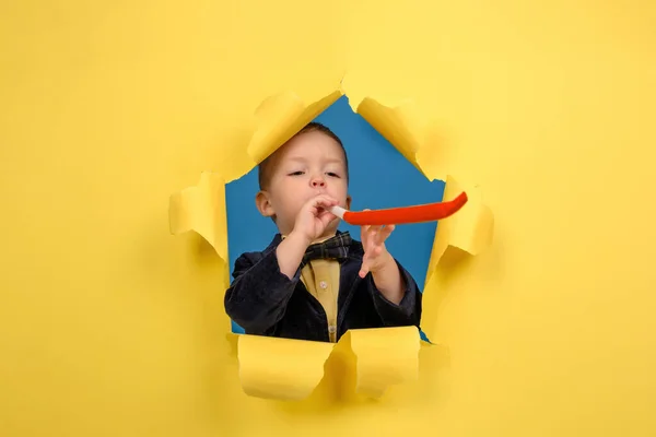 Aniversariante menino de jaqueta e gravata borboleta sopra apito vermelho festivo no fundo do estúdio amarelo rasgado. Concepção de um feriado, bom humor e aniversário. — Fotografia de Stock