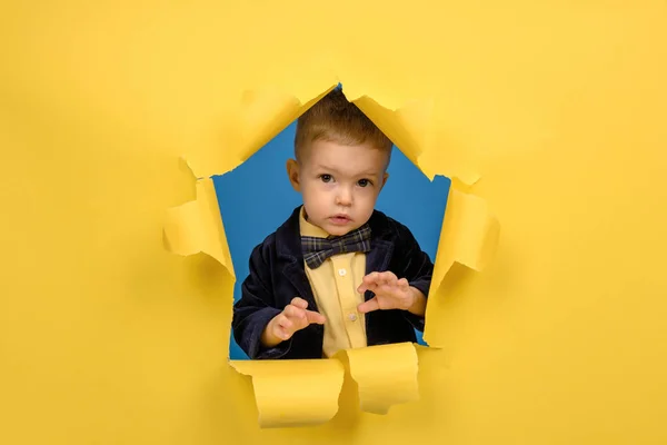Boy looks out of hole of torn yellow paper with torn edges in jacket and bow tie and shows good positive emotion on his face. Concept of an advertising poster and good holiday mood with place for text — Stock Photo, Image