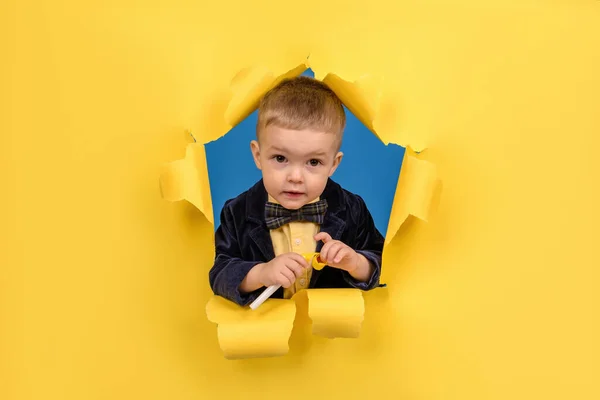 Happy birthday boy in jacket and bow tie holds yellow holiday whistle on ragged yellow Studio background. Concept of a holiday, good mood and birthday. — Stock Photo, Image
