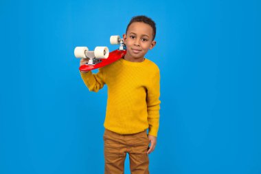 Sıradan giyinmiş, sarı kazaklı, kahverengi pantolonlu, kırmızı kaykaylı ve mavi arka planda gülümseyen sevimli Afro-Amerikalı ergen. Aktivite ve mutlu çocukluk kavramı